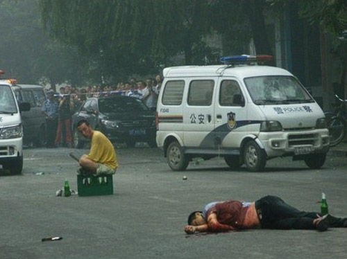In Liaoning, China, a policeman lies on the street having been stabbed to death while his assailant sits on a beer crate at a distance.