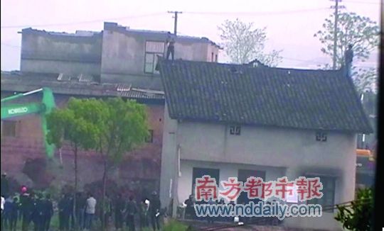 Chinese man standing on roof with bottle of gasoline as an excavator approaches to tear down his house.