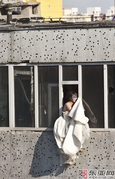 A young Chinese girl in a wedding dress hangs out of a window, distraught, thinking of suicide.