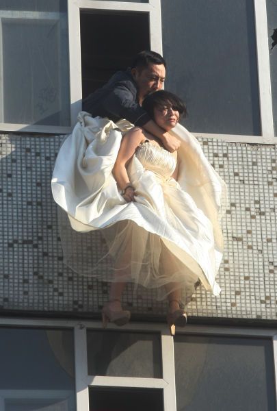 A Chinese girl in a wedding dress hangs in mid-air, a man leaning out of a window struggling to hold onto her around her neck.